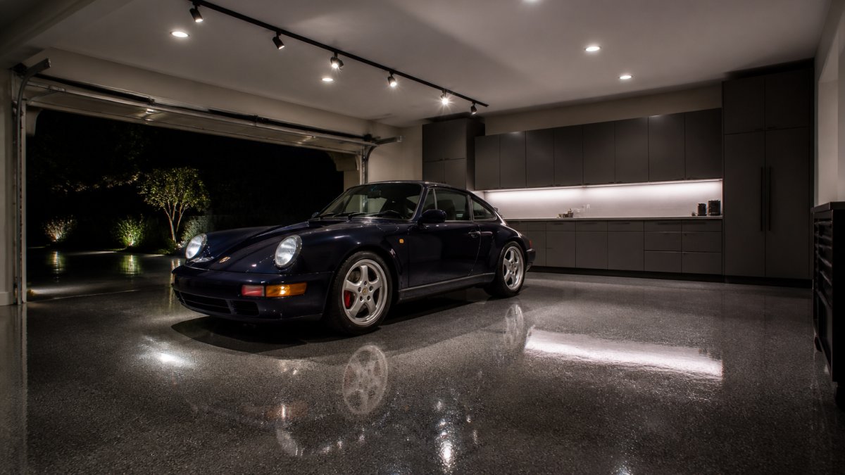A collector's garage showroom at night with track lighting and recessed ceiling lights illuminating a classic sports car, glossy epoxy floor reflecting the light, custom cabinetry along the wall